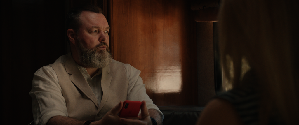 Der Protagonist des Films, Chuck, sitzt in seinem Wohnmobil mit seinem Handy in der Hand, im gegenüber sitzt die Roboterfrau Harmony. 