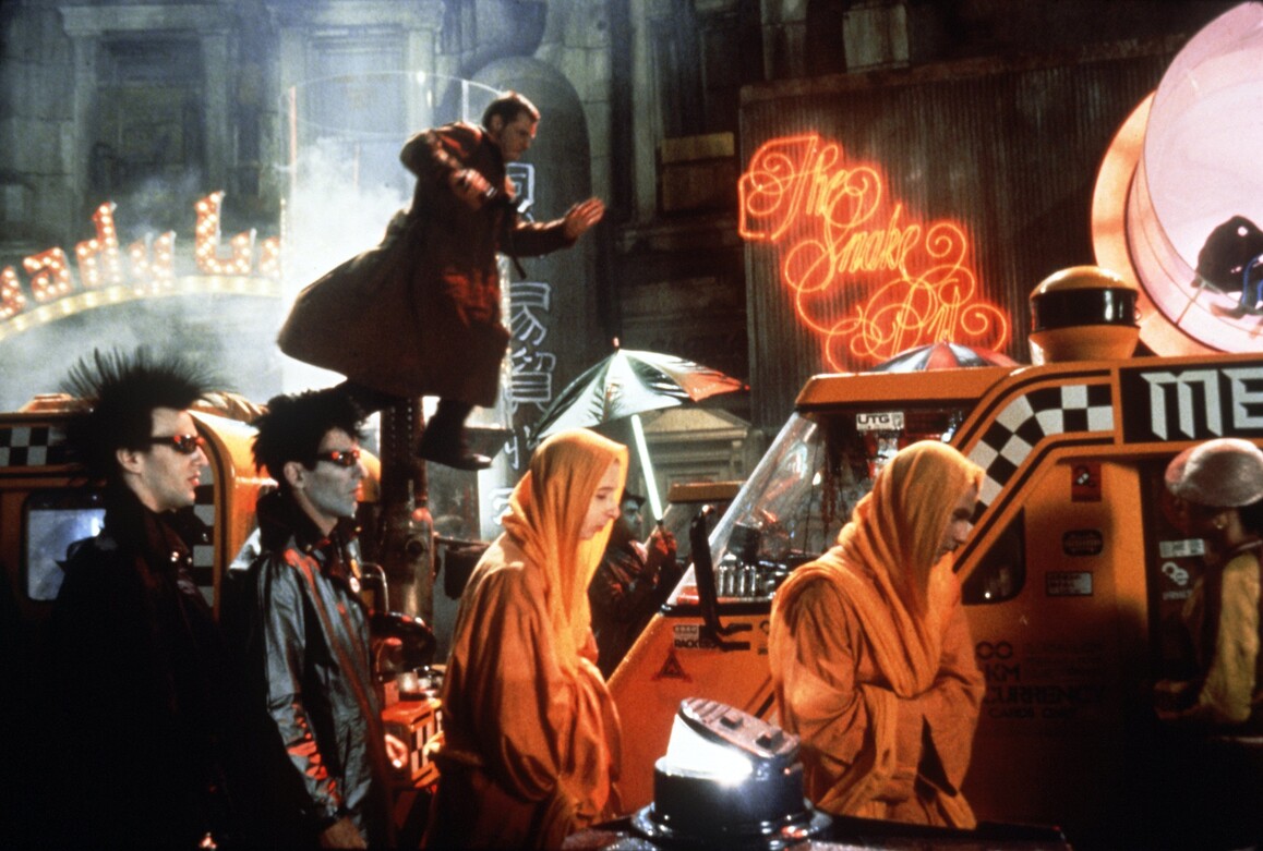 Eine düstere Straßenszenerie ist dargestellt, es ist nachts, im Vordergrund laufen vermummte Gestalten, im Hintergrund sieht man den Replikantenjäger Deckard. 