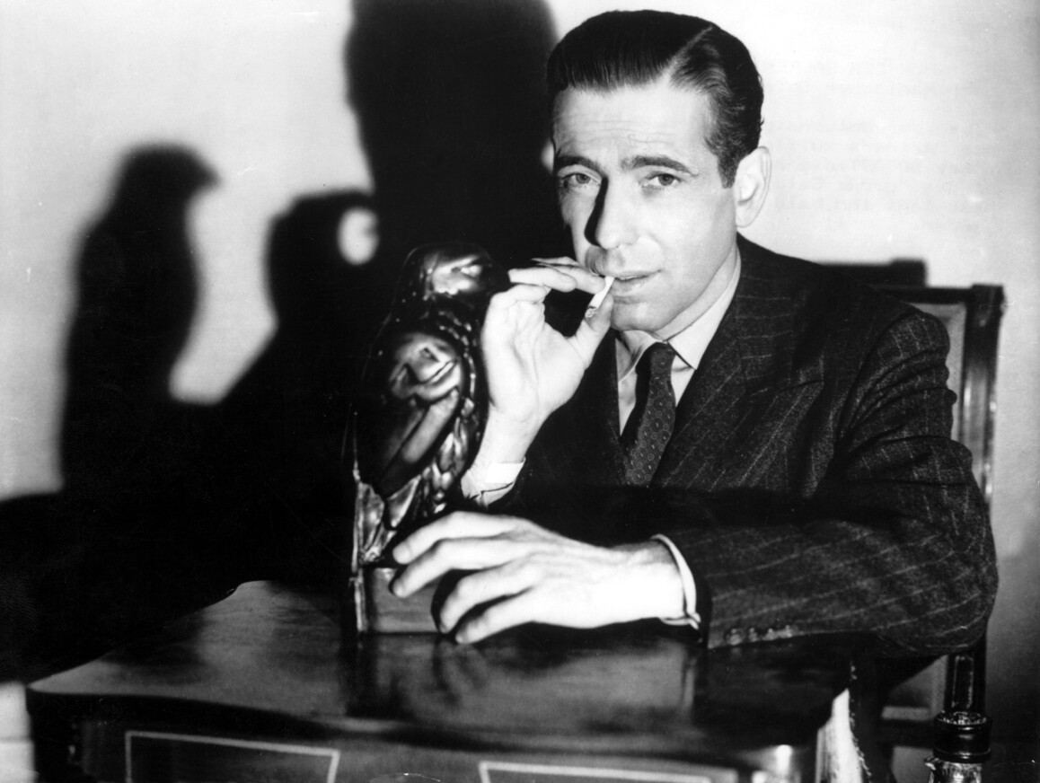 Auf der Fotografie ist Humphrey Bogart dargestellt. Er zieht an einer Zigarette und blickt direkt in die Kamera.