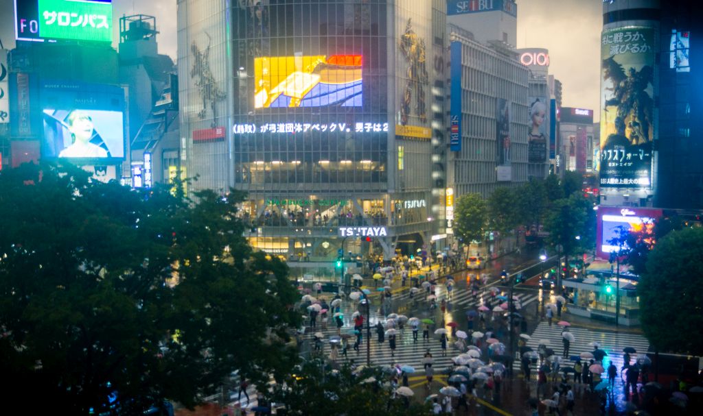 Eine nächtliche Stadtszenerie aus Tokio ist zu sehen, Hochhäuser, Leuchtreklamen und viele Menschen sind abgebildet. 